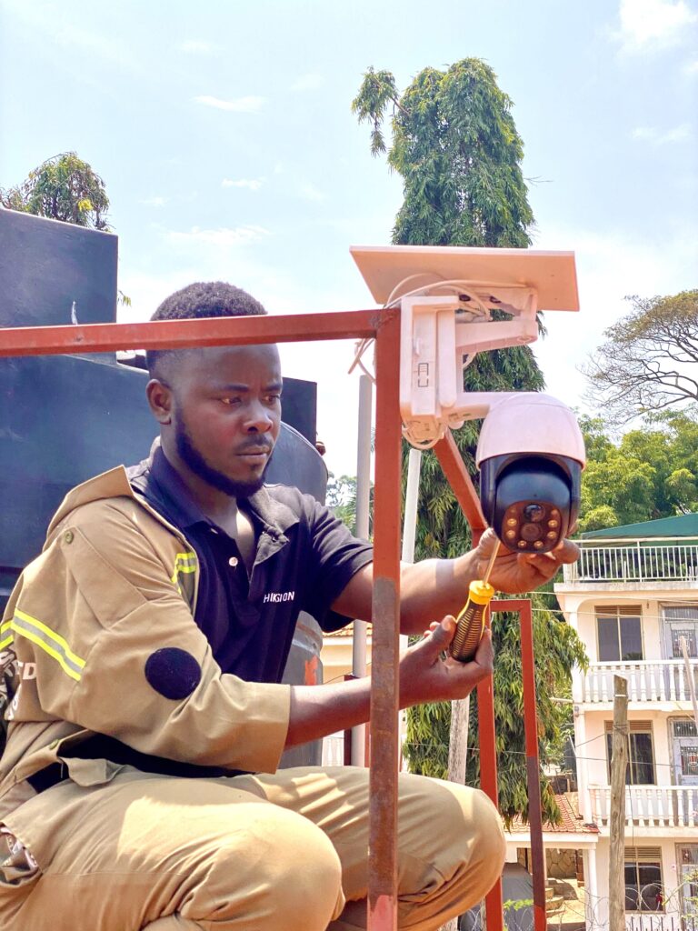 A high-tech solar-powered CCTV camera mounted on a steel pole overlooking a construction site in Wakiso, Uganda, featuring a large solar panel and 4G antenna.